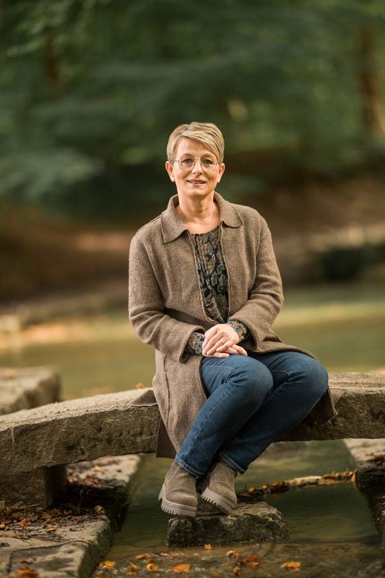 Maren Frank sitz auf einer kleinen Steinbrücke, die über einen kleinen Wasserlauf führt. Sie lächelt in die Kamera.