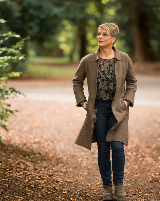 Maren Frank geht auf einem von Laub bedeckten Weg durch einen Park oder Wald und trägt einen braunen Mantel.