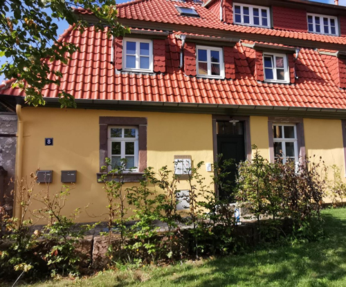 Gelbes Gebäude mit roten Dachziegeln bei Sonnenschein. An der Hauswand hängen Briefkästen und die Hausnummer 8.